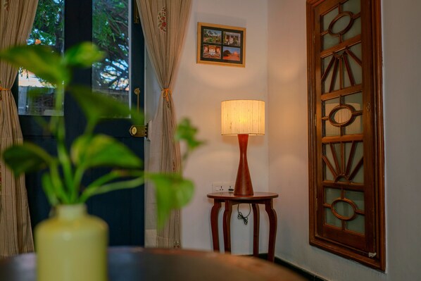 A room with a wooden door, a small table with a lamp, and a plant in the foreground. - Residence De L'eveche - CGH Earth (Puducherry)
