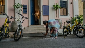 Entrada de um edifício com uma porta azul, duas bicicletas estacionadas do lado de fora e uma pessoa inclinada no chão.