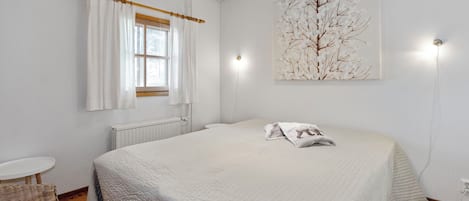 Un dormitorio con cama, ventana con cortinas, luz empotrada en la pared, radiador, una mesita y una silla.
