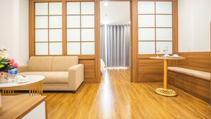 Ein Zimmer mit Holzboden, einem beigen Sofa, einem runden Tisch mit weißer Tischdecke und einem kleinen Tisch mit Blumen.