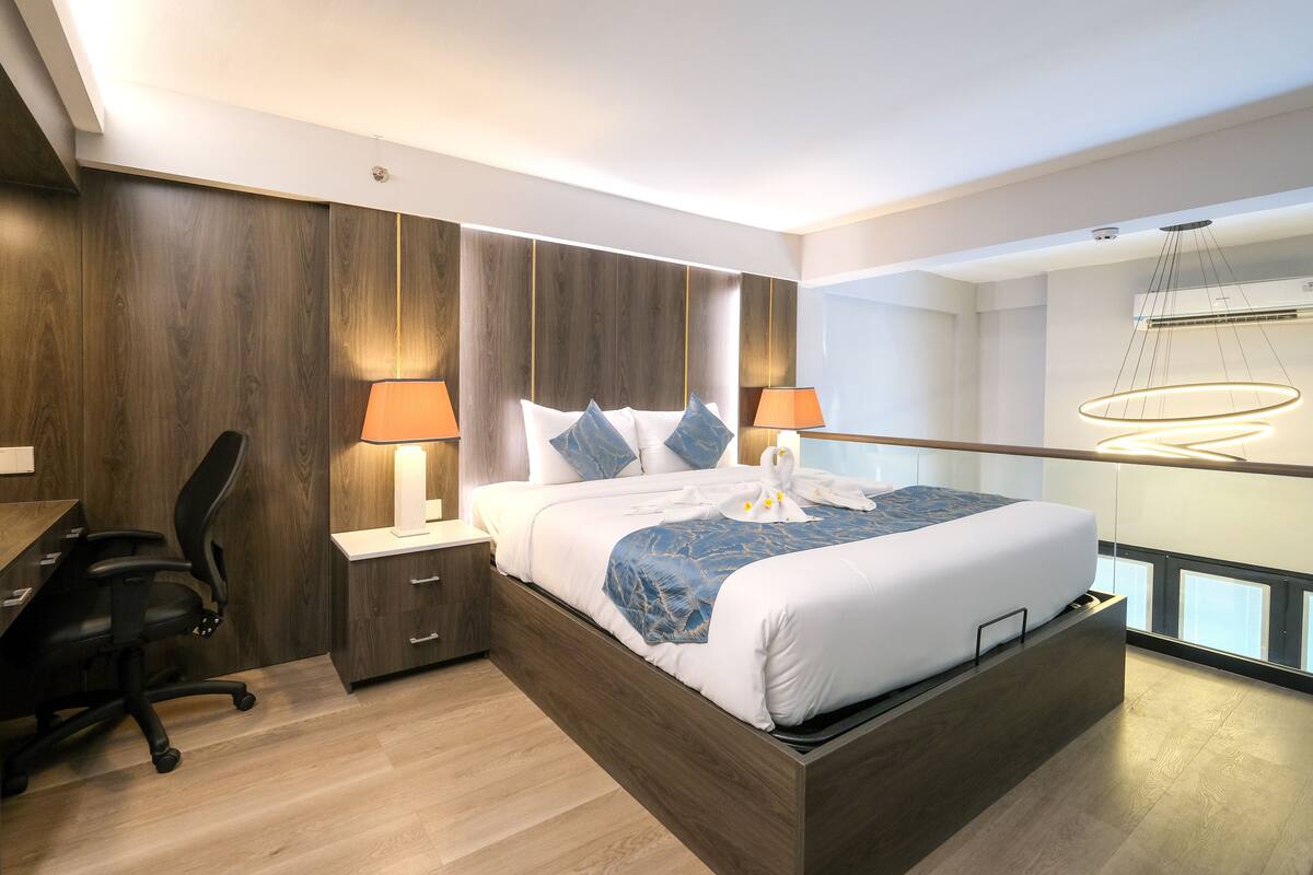 A modern hotel room with a wooden headboard, a large bed with white bedding, two bedside lamps, a desk with a chair, and a view of the city through a large window.