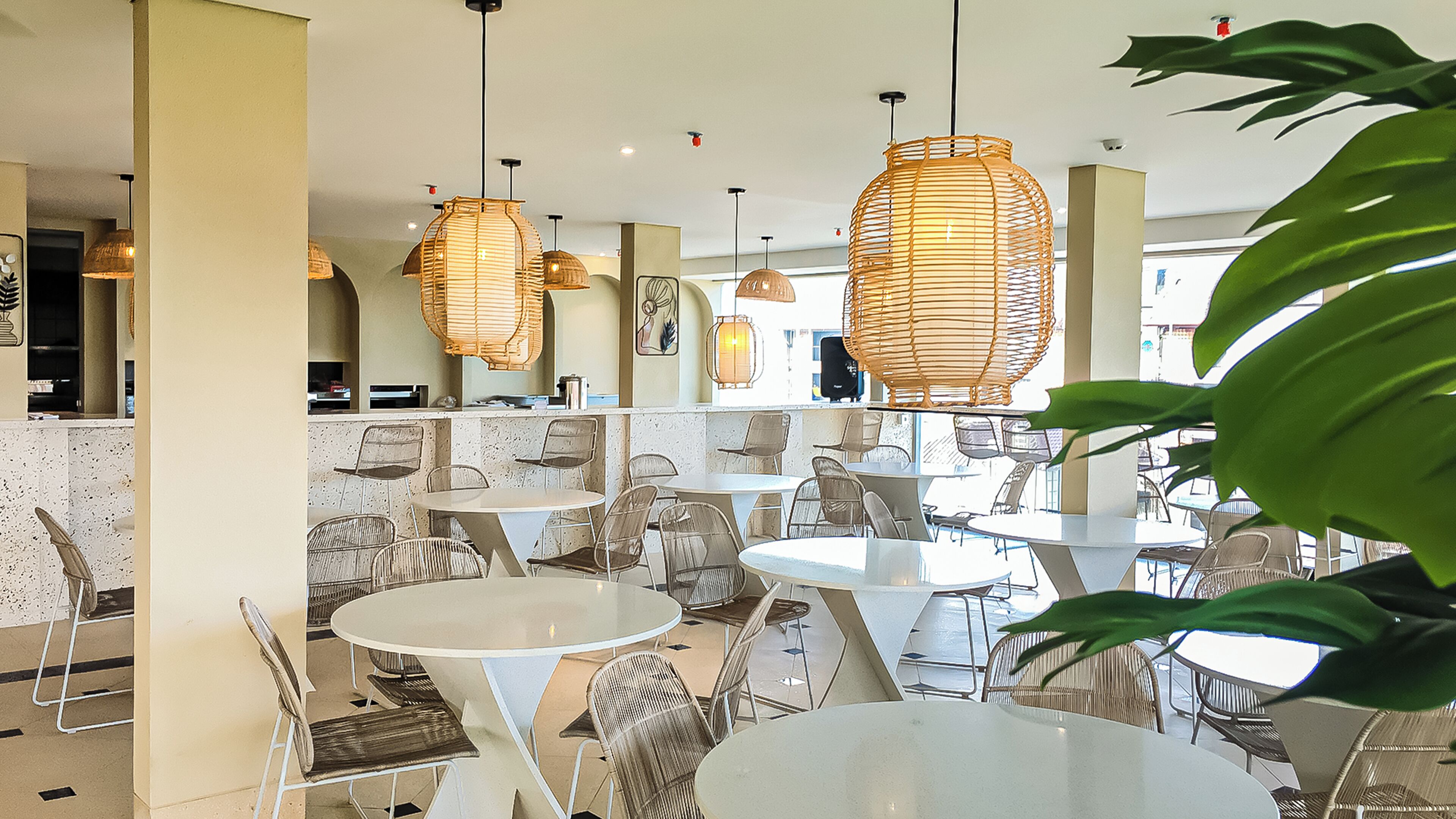 L’intérieur d’un restaurant, avec des tables rondes et des chaises en osier, agrémenté de lampes en osier suspendues et offrant une vue sur la rue.