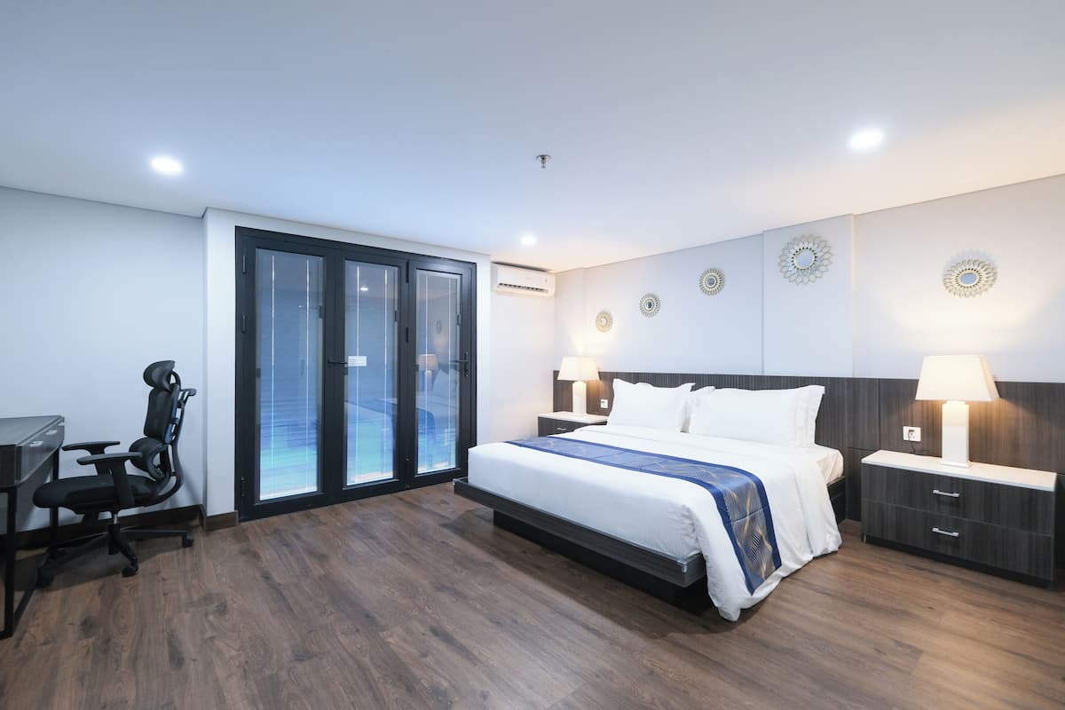A modern hotel room with a large bed, a desk, and a chair. There are decorative wall sconces and a view through the glass door.