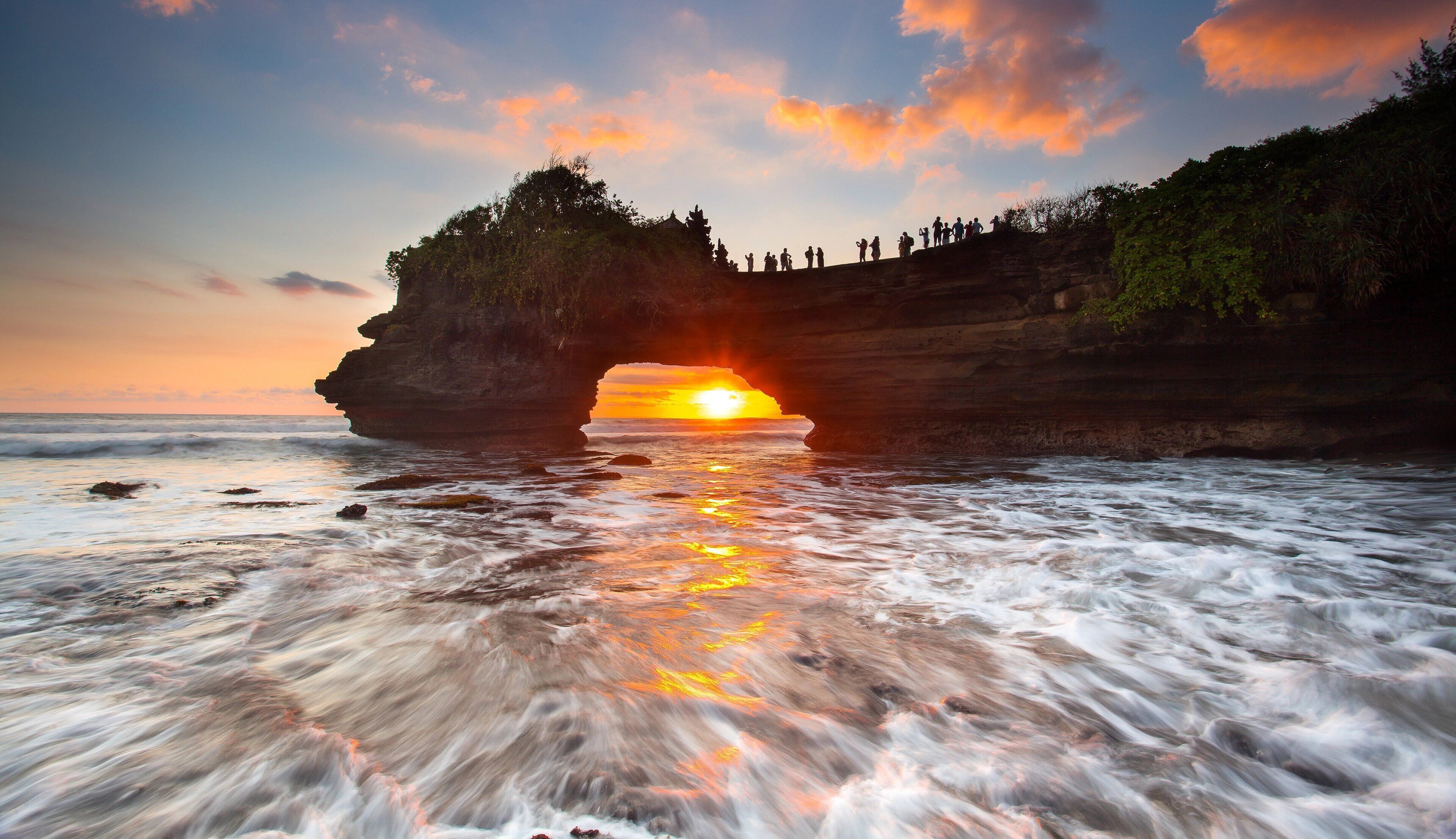 A natural rock formation with a cave entrance, a sunset, and a group of people at the top.