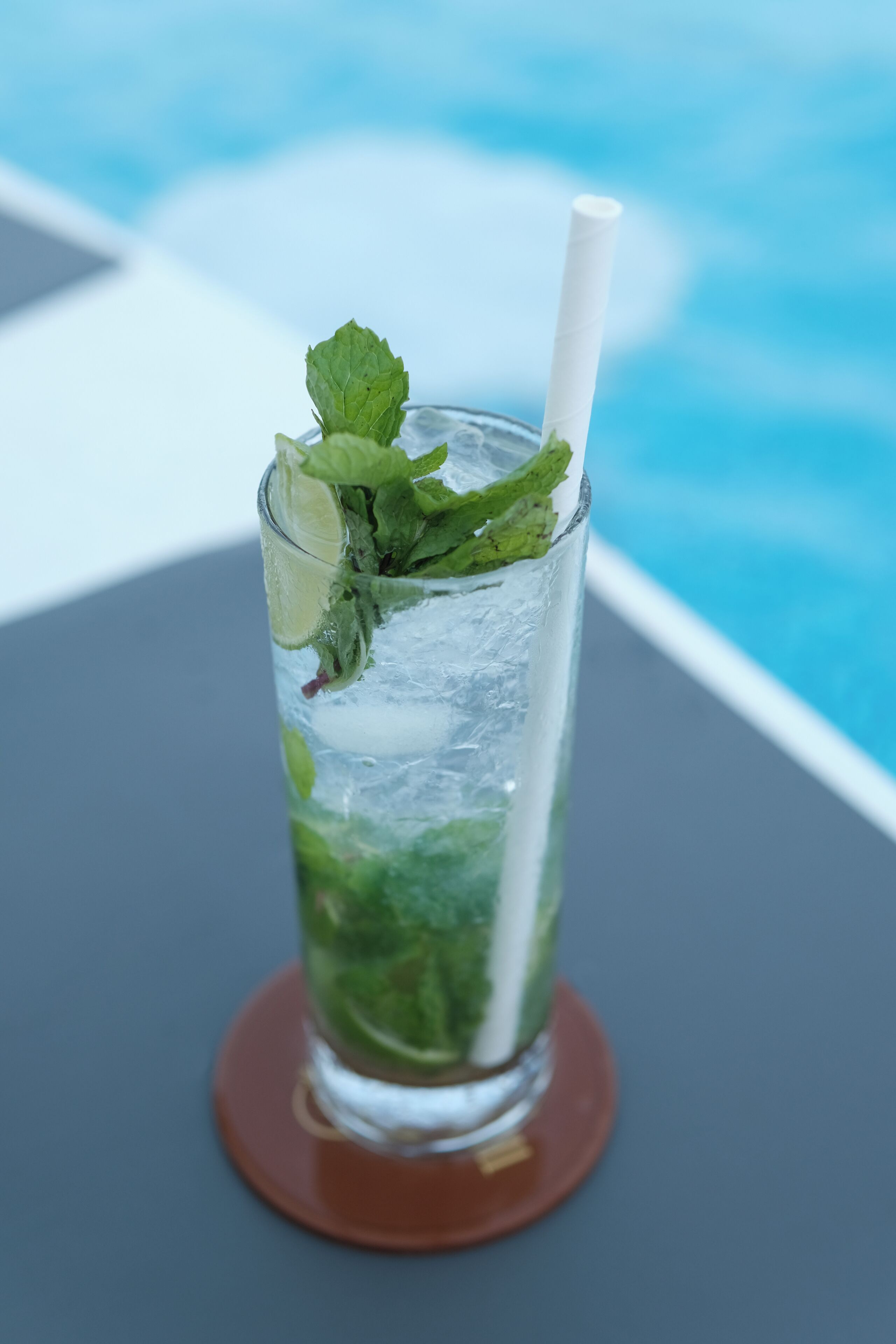 A glass of iced beverage with mint leaves and a straw, placed on a table with a pool view in the background.