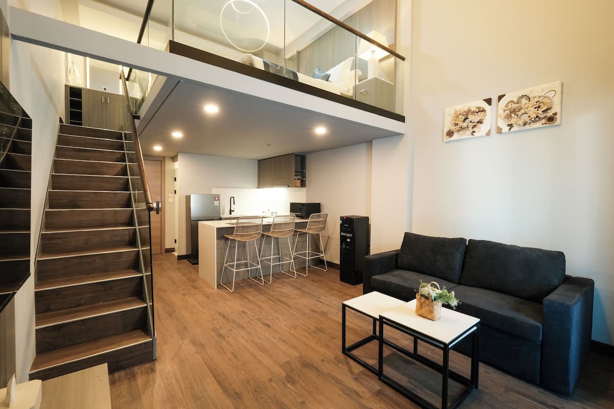 A modern interior with a staircase, a kitchen area, a living room with a sofa, and two artworks on the wall.