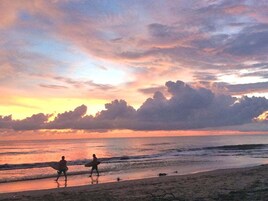 Escena de playa con dos personas caminando por la orilla, llevando tablas de surf.