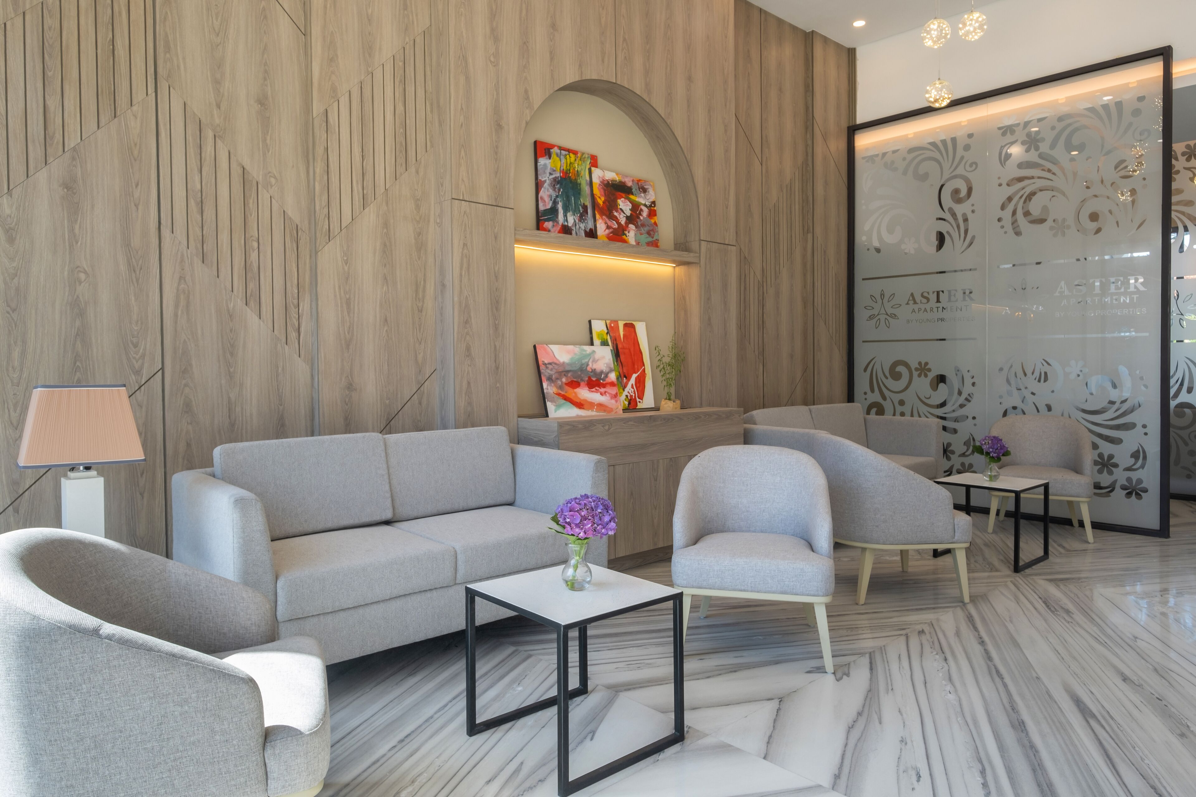Un moderno vestíbulo con un sofá gris, sillones, una mesita con un florero y un mostrador de recepción con obras de arte.