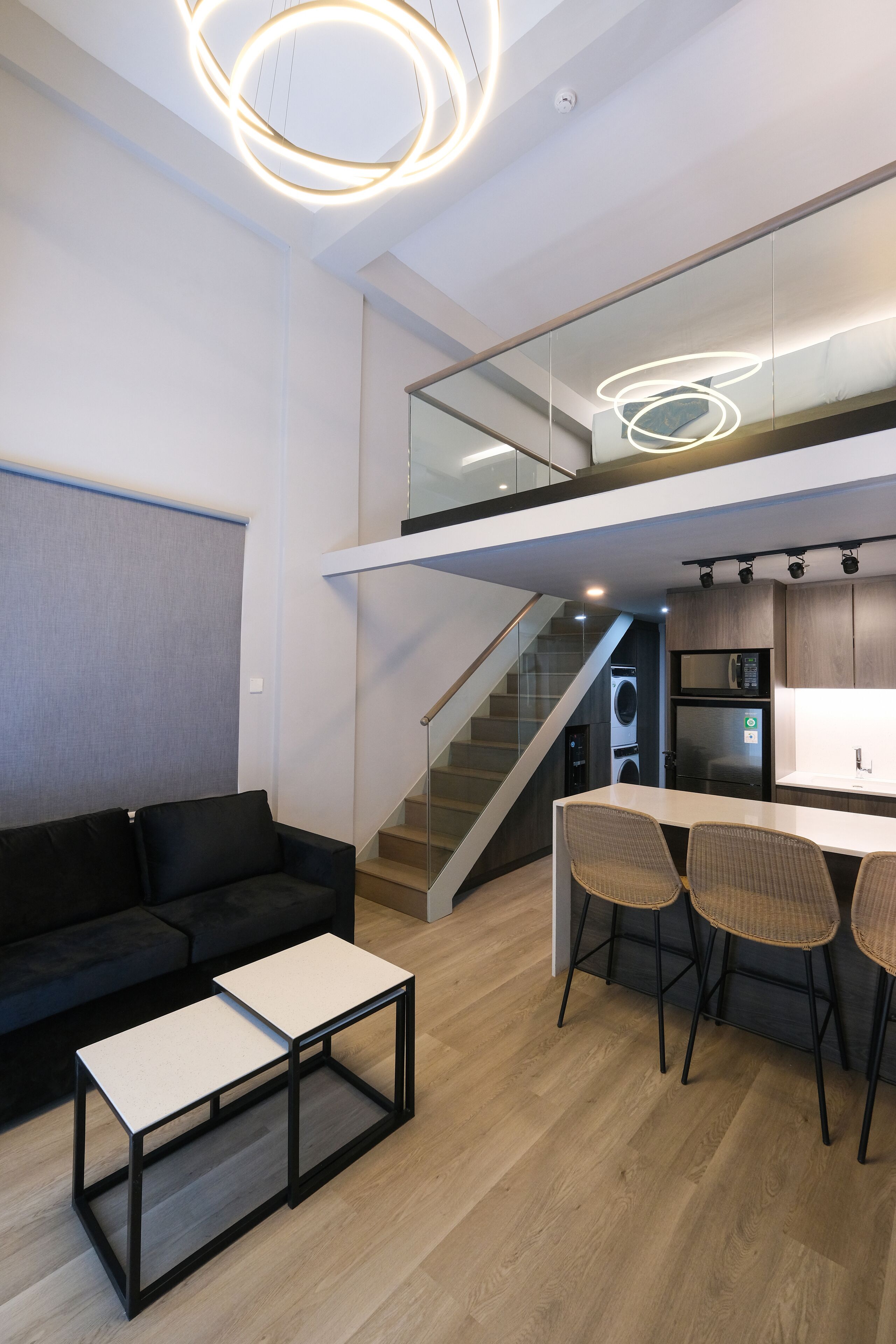 A modern kitchen with a central island, a dining area with chairs, a staircase leading to an upper level, and a black sofa in the living area.
