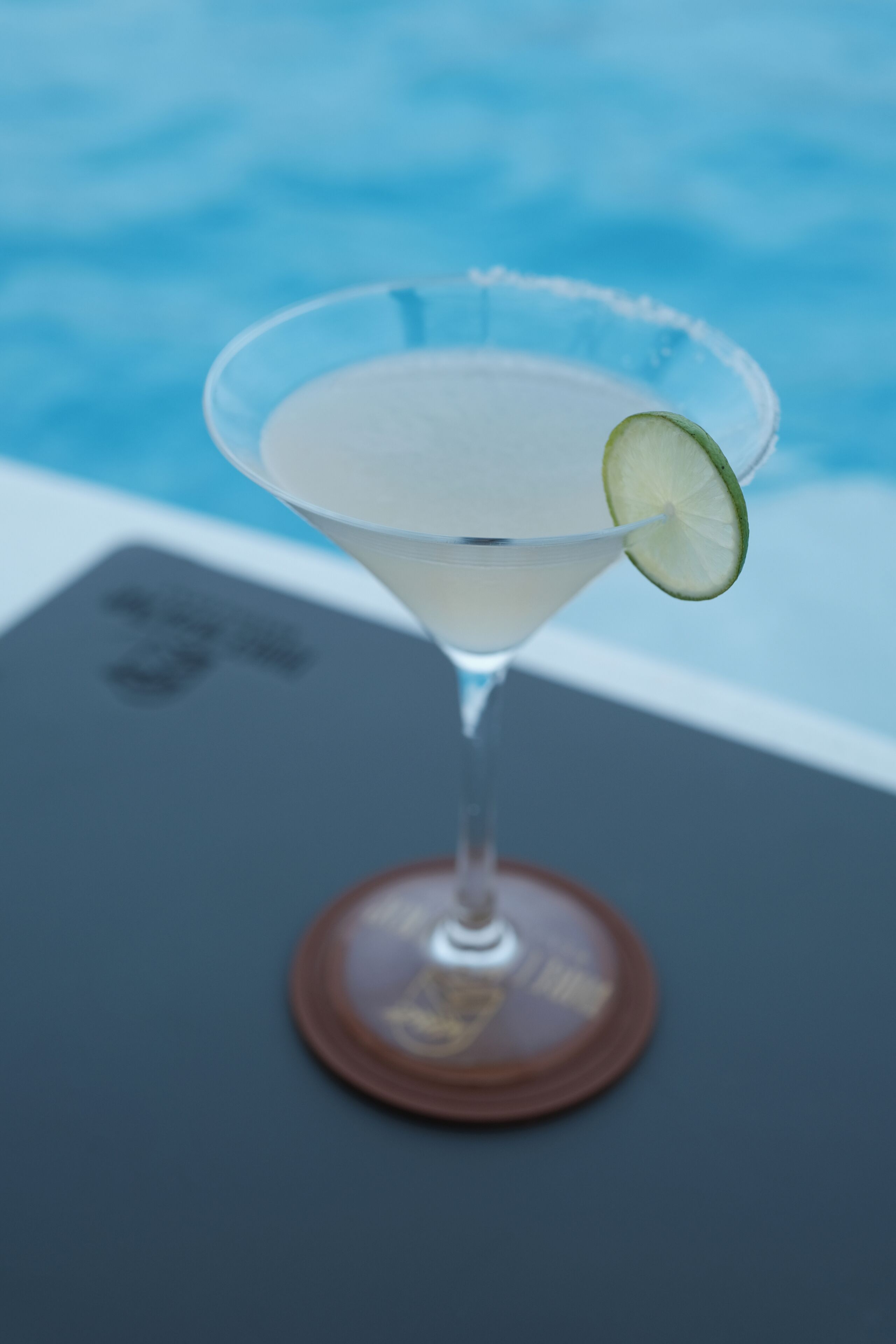 Un verre à martini, garni d’une tranche de citron vert, est posé sur une table au bord d’une piscine.