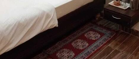 A single bed with a dark headboard and two pillows, a small bedside table with a kettle, and a patterned rug on a wooden floor.