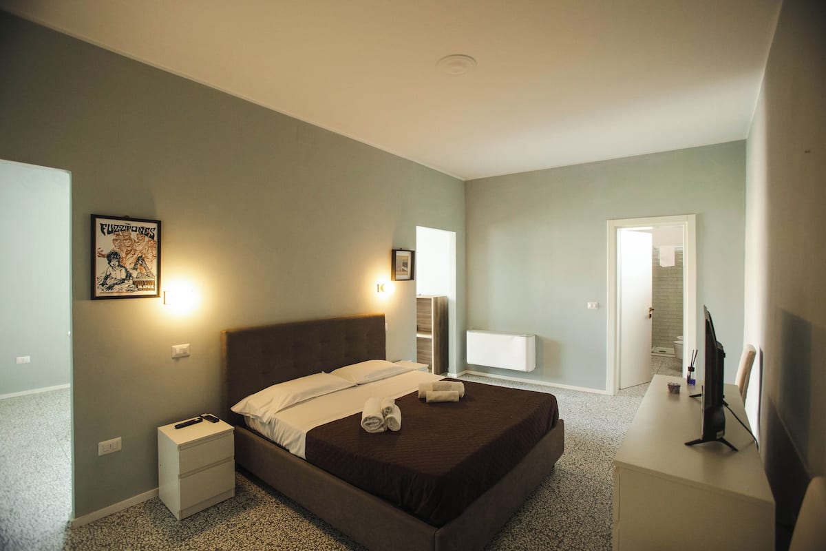 Une chambre d’hôtel bien rangée, avec un lit, des tables de chevet, un bureau avec un ordinateur et une salle de bain visible à travers une porte ouverte.