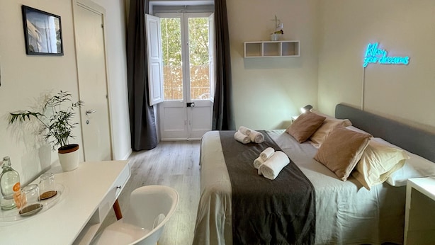 A modern bedroom with a bed, bedside tables, a desk, a chair, a potted plant, and a window with curtains. - 4Brooms-Paladini b&b (Lecce)
