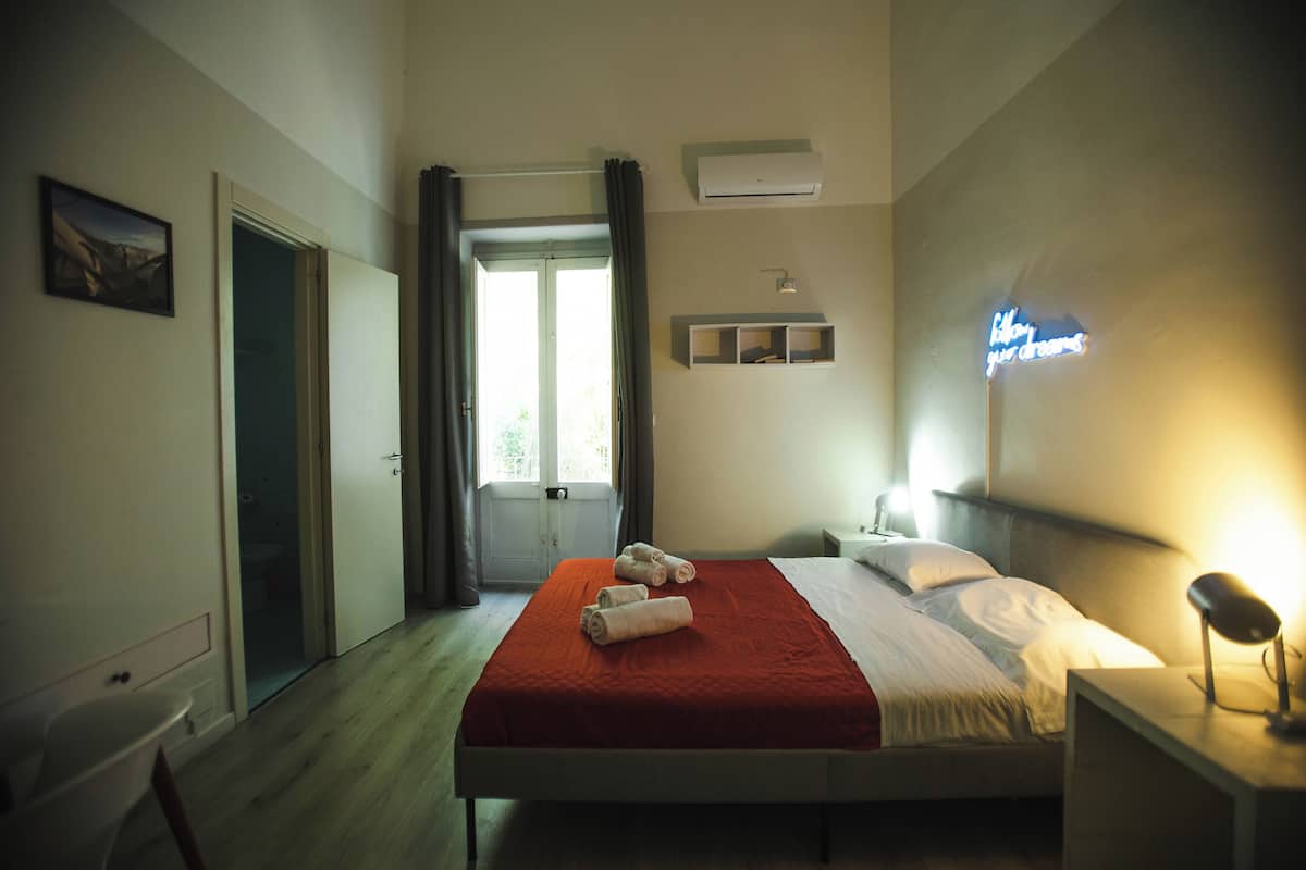 Une chambre à coucher comprenant un lit, une table de chevet, une commode, une étagère fixée au mur, une porte et une fenêtre avec des rideaux.