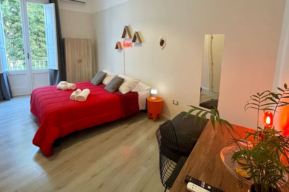 Une chambre moderne avec un lit rouge, un bureau en bois, une chaise, une plante en pot et une lampe de chevet.
