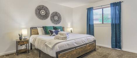 A bedroom with a bed, two wall decor pieces, a nightstand, a lamp, and a window with curtains.