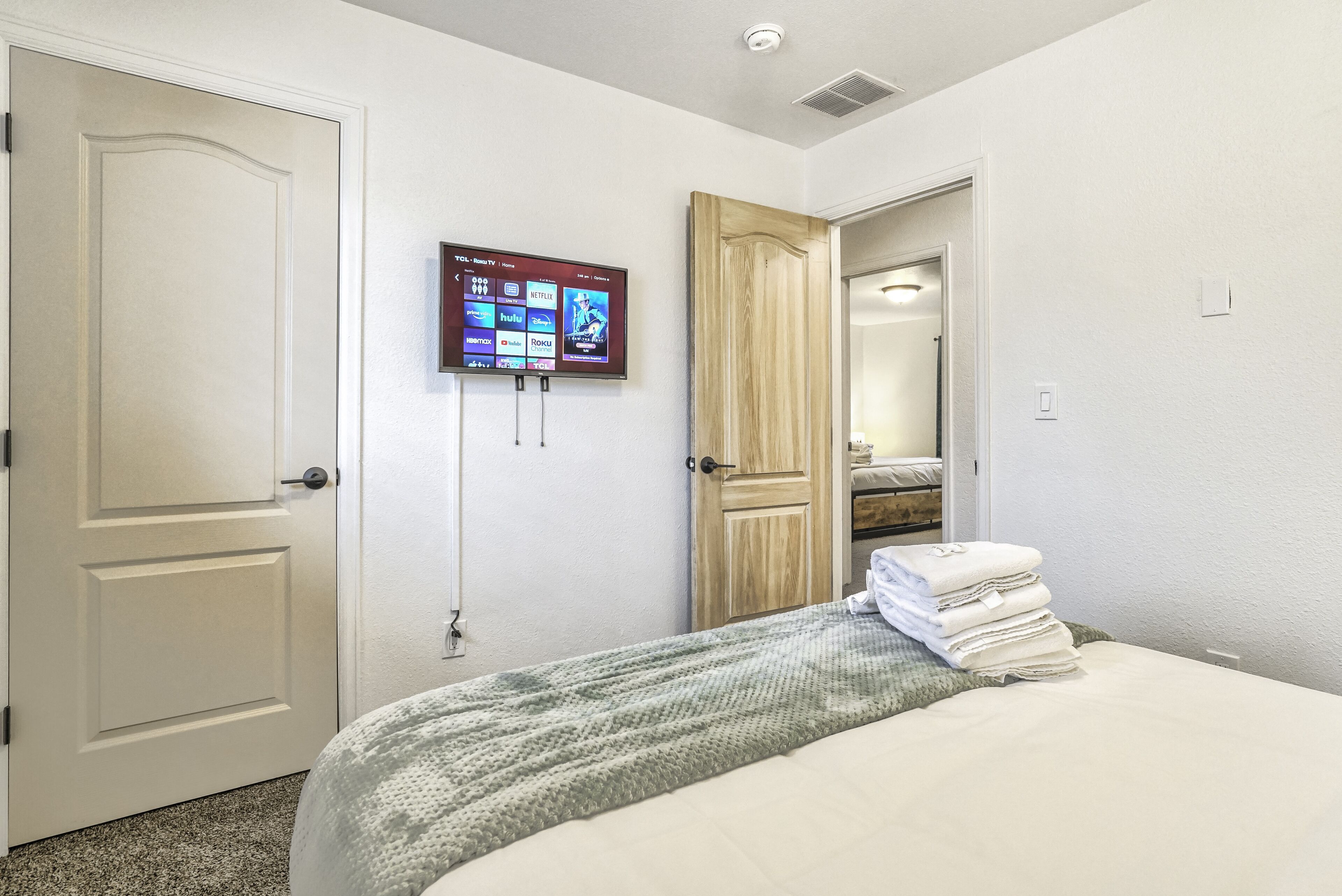 A bedroom with a bed, a TV mounted on the wall, and a door leading to another room.