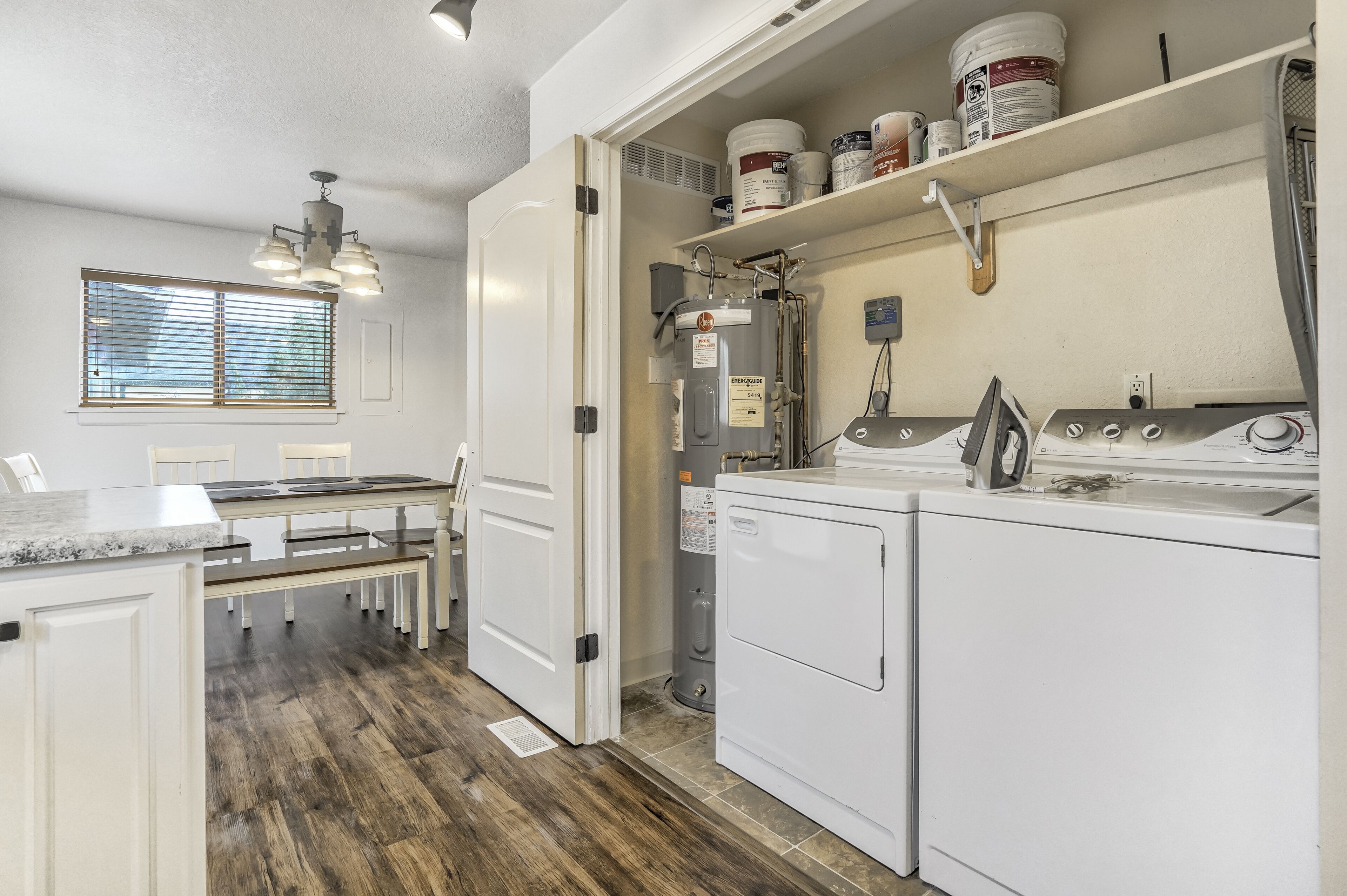 A laundry room with a washing machine, dryer, and water heater. There is a window with blinds, a bench, and a countertop with a sink.