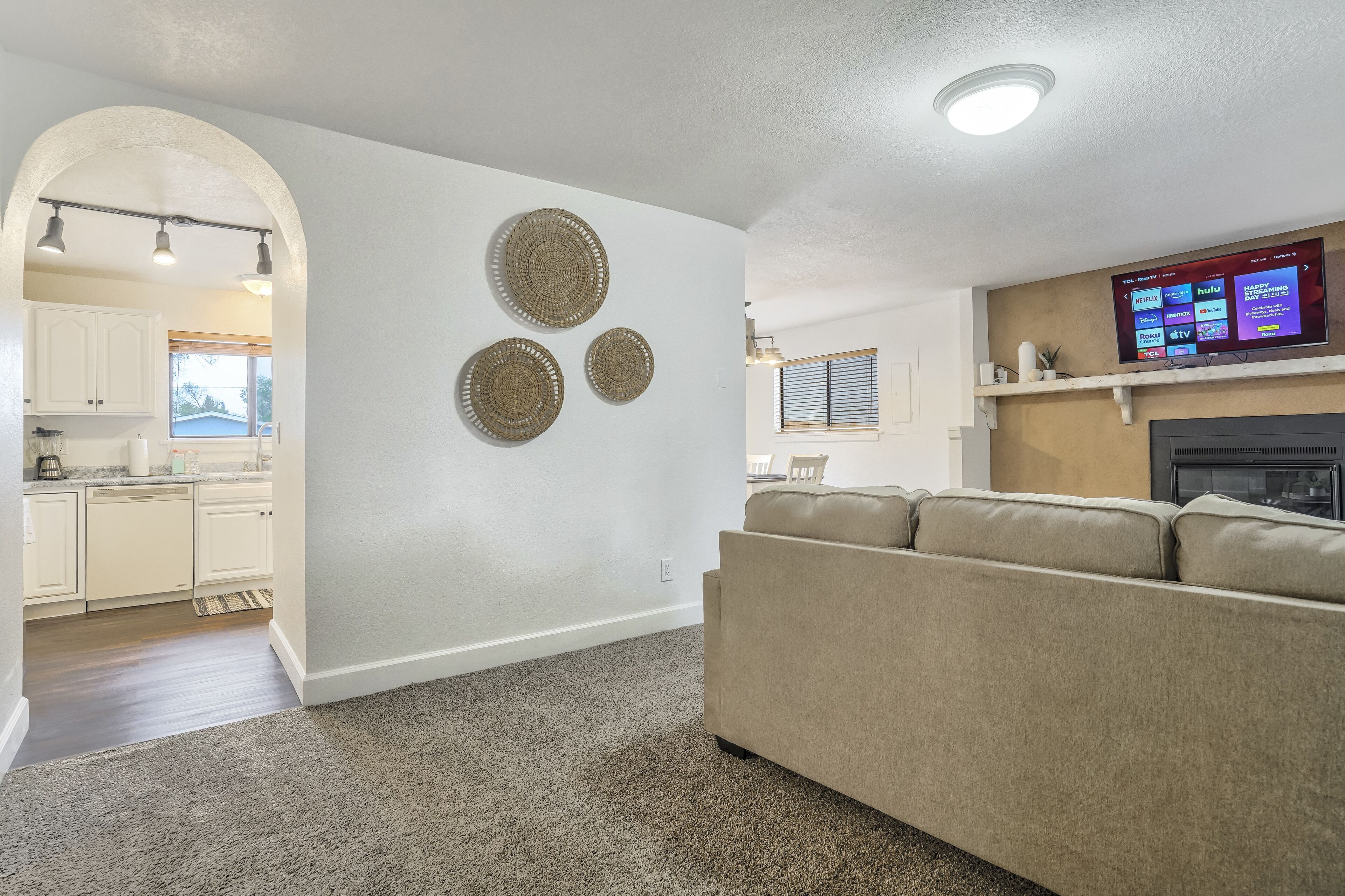 A living room with a beige sofa, a flat-screen TV mounted on the wall, and a kitchen visible through an archway.