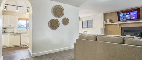 A living room with a beige sofa, a flat-screen TV mounted on the wall, and a kitchen visible through an archway.