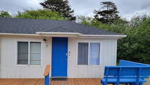A small, single-story building with a blue door and windows, a wooden deck, and a blue bench. - Outstanding Location, Pet Friendly, Kitchen, WiFi, Two Blocks From Beach, Unit 7 (Rockaway Beach)