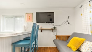 A small, cozy living room with a grey sofa, a blue chair, a yellow cushion, a TV mounted on the wall, and a window with blinds. - Outstanding Location, Pet Friendly, Kitchen, WiFi, Two Blocks From Beach, Unit 7 (Rockaway Beach)