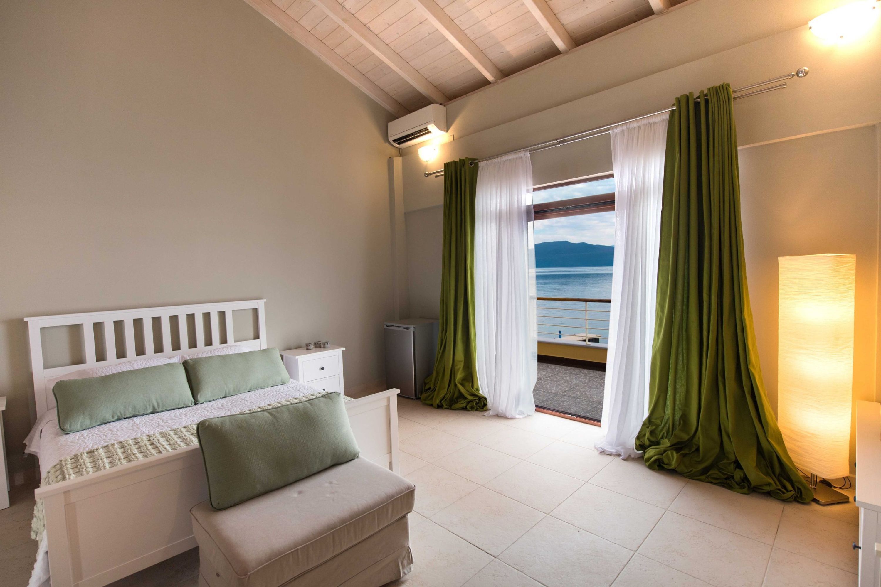 Une chambre à coucher avec un lit, une table de chevet, une lampe et une vue sur la mer, visible à travers les rideaux.