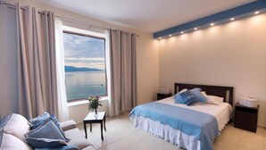A modern bedroom with a large window offering a view of the sea, a bed with blue bedding, a sofa with blue cushions, and a small table with a vase of flowers. - NV Seafront Studios AV Properties (Lefkada)