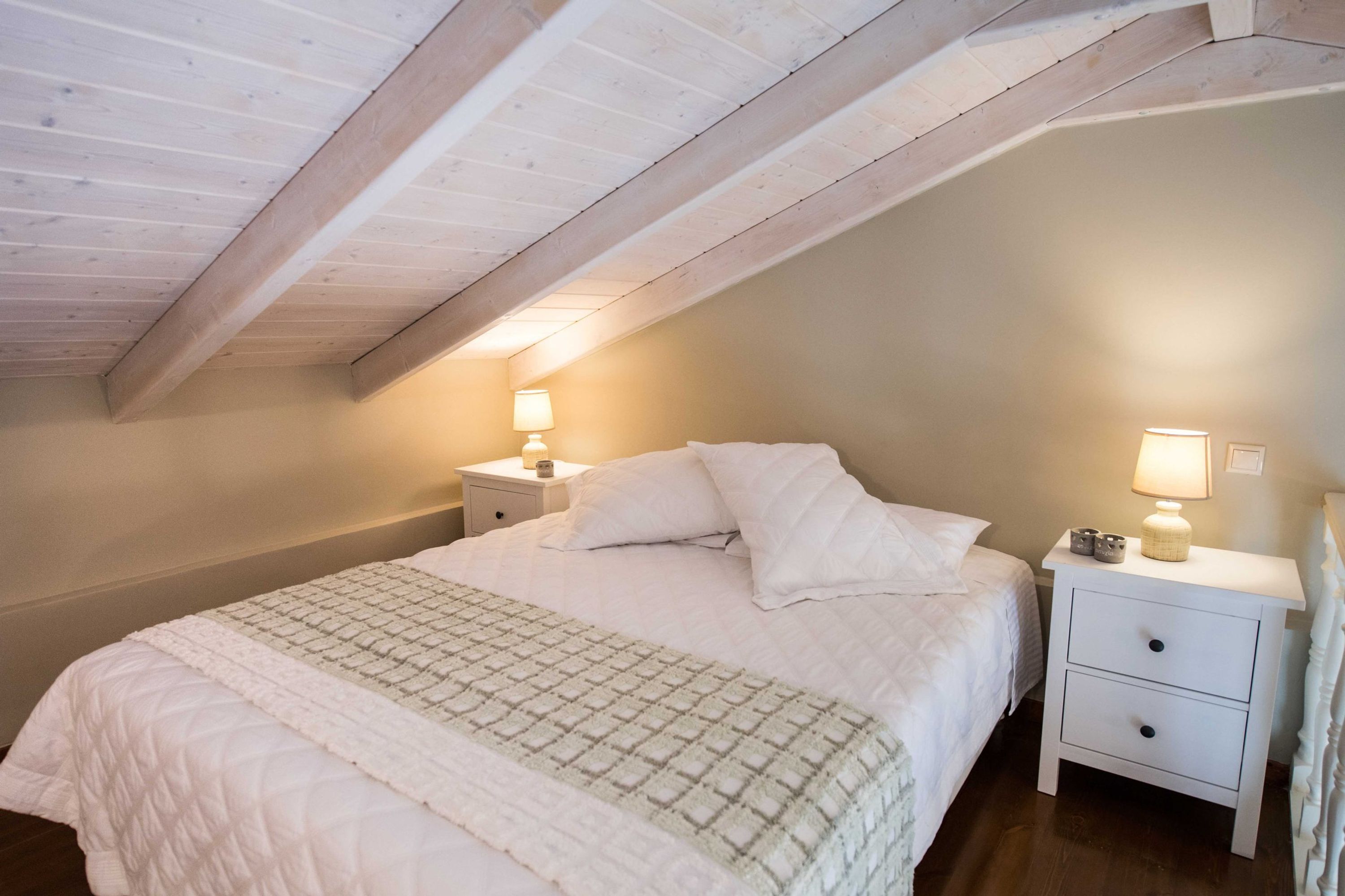 Un dormitorio con techo alto, una cama con ropa blanca, dos mesitas de noche con lámparas y una viga de madera en el techo.