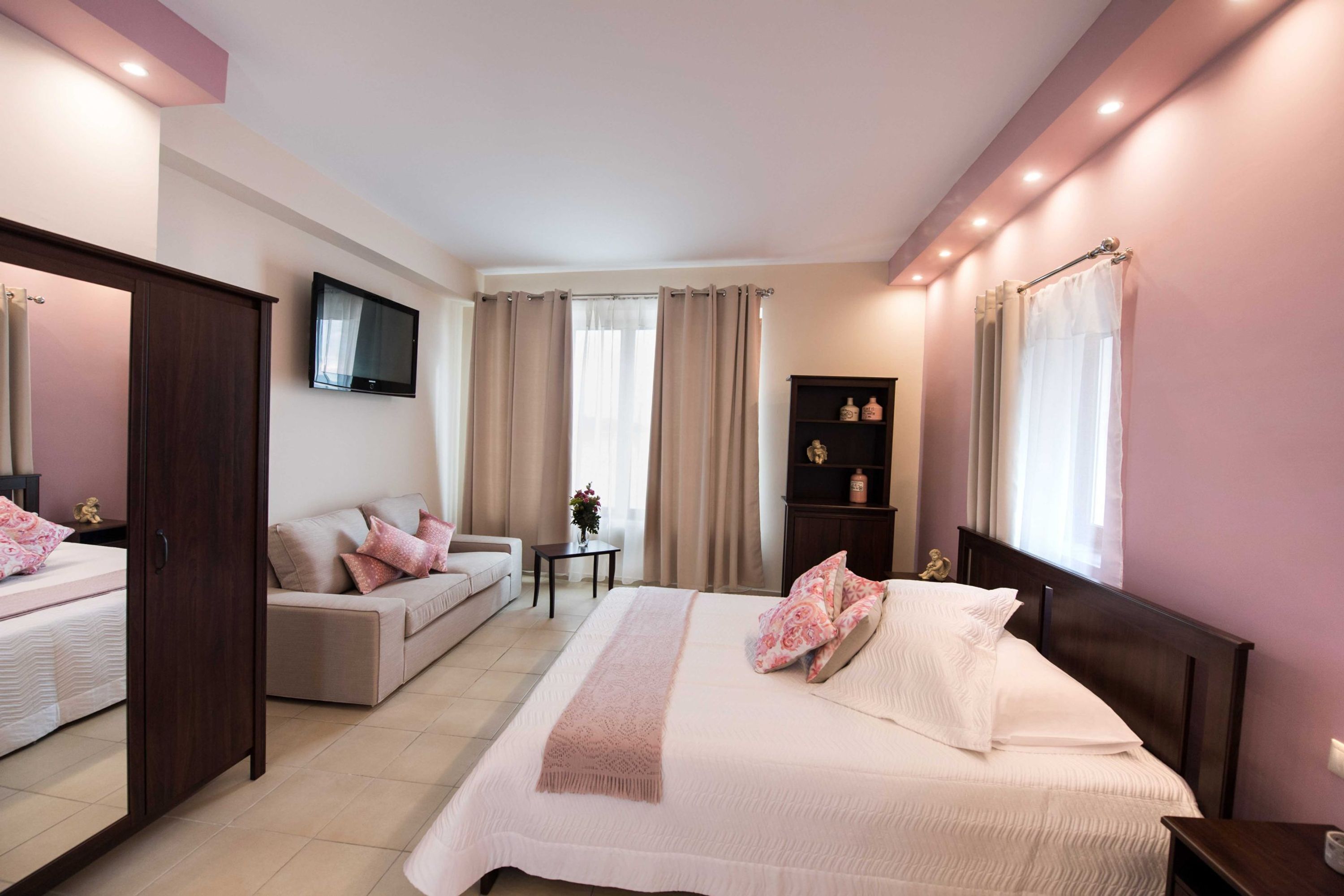A bedroom with a bed, a sofa, a TV, a small table with a vase, and a shelf with bottles.