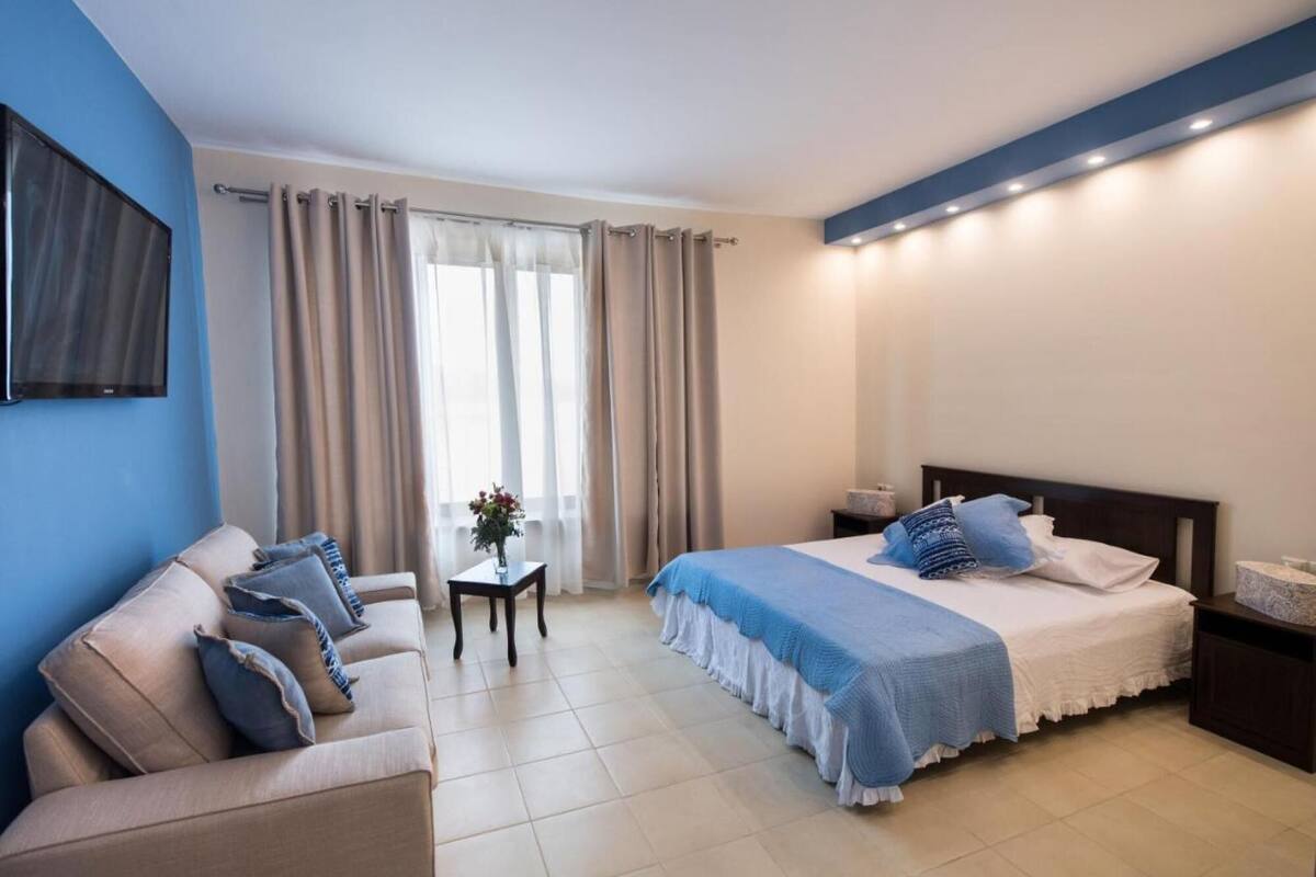 A hotel room with a bed, a sofa, a small table with a vase, and a TV mounted on the wall.
