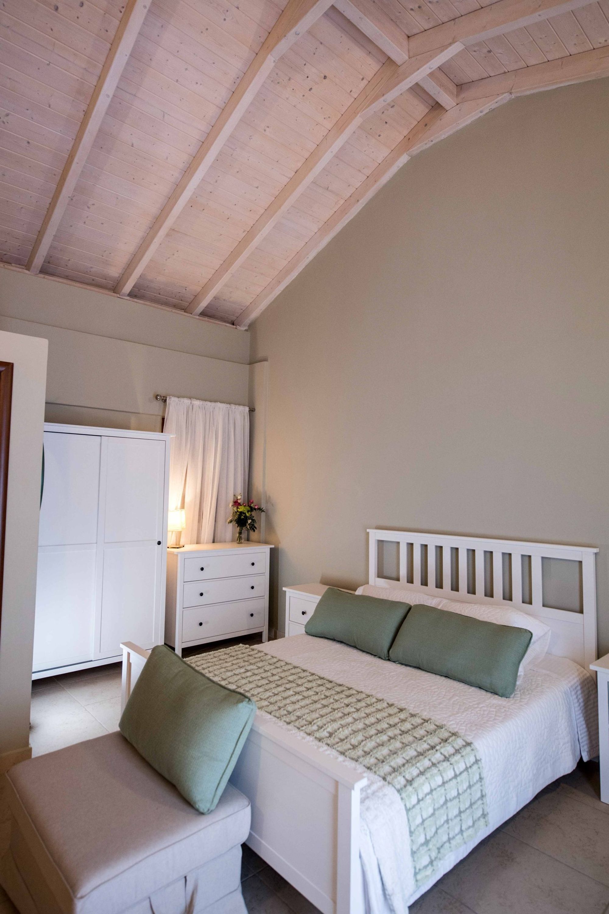 A bedroom with a wooden ceiling, a bed with green pillows, a nightstand, a dresser, and a chair.