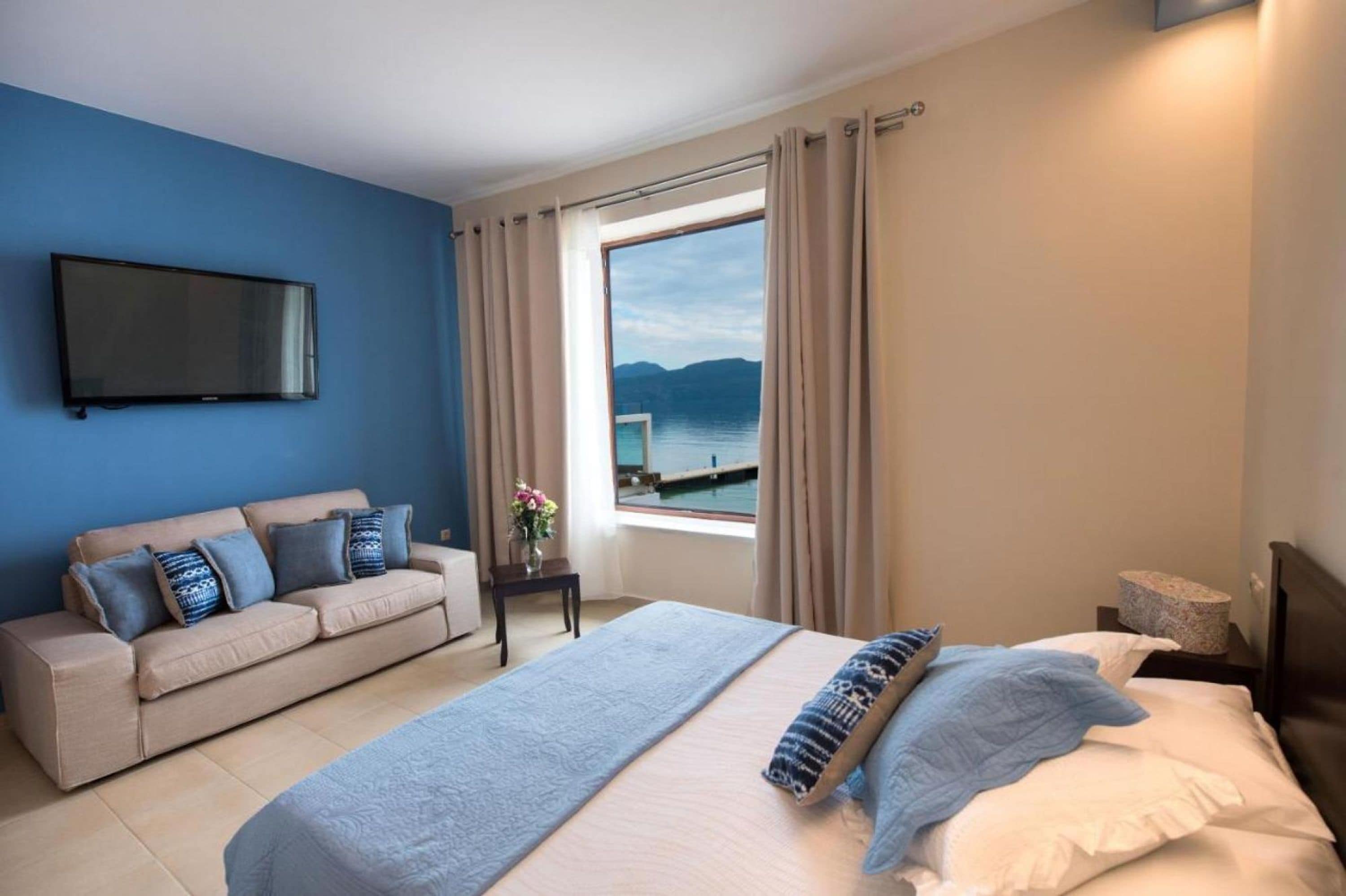 A hotel room with a bed, a sofa, a TV, and a window with a view of the sea.