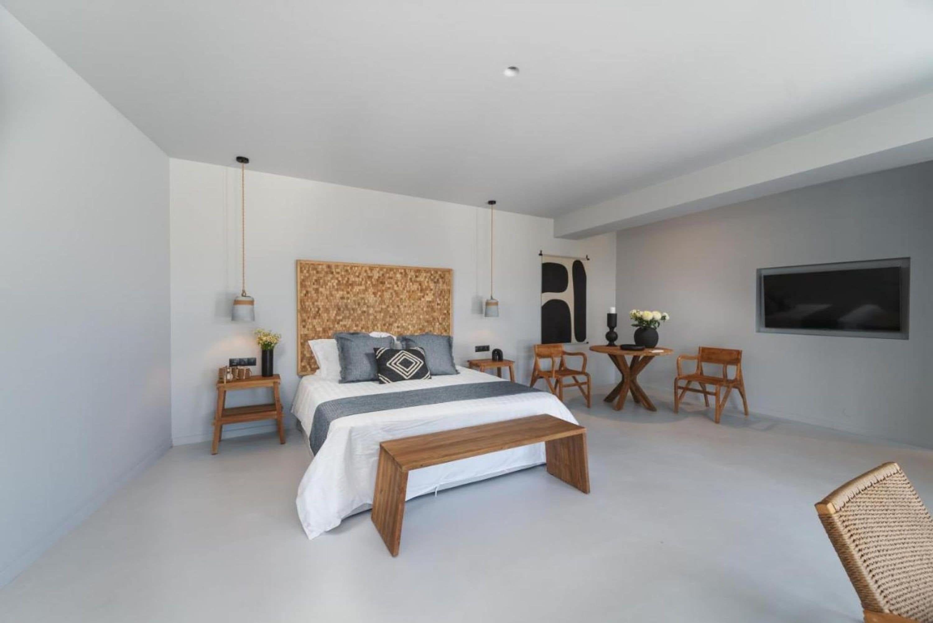 Une chambre moderne avec un grand lit, des tables de chevet, un banc en bois, une petite table ronde avec des chaises et un téléviseur fixé au mur.