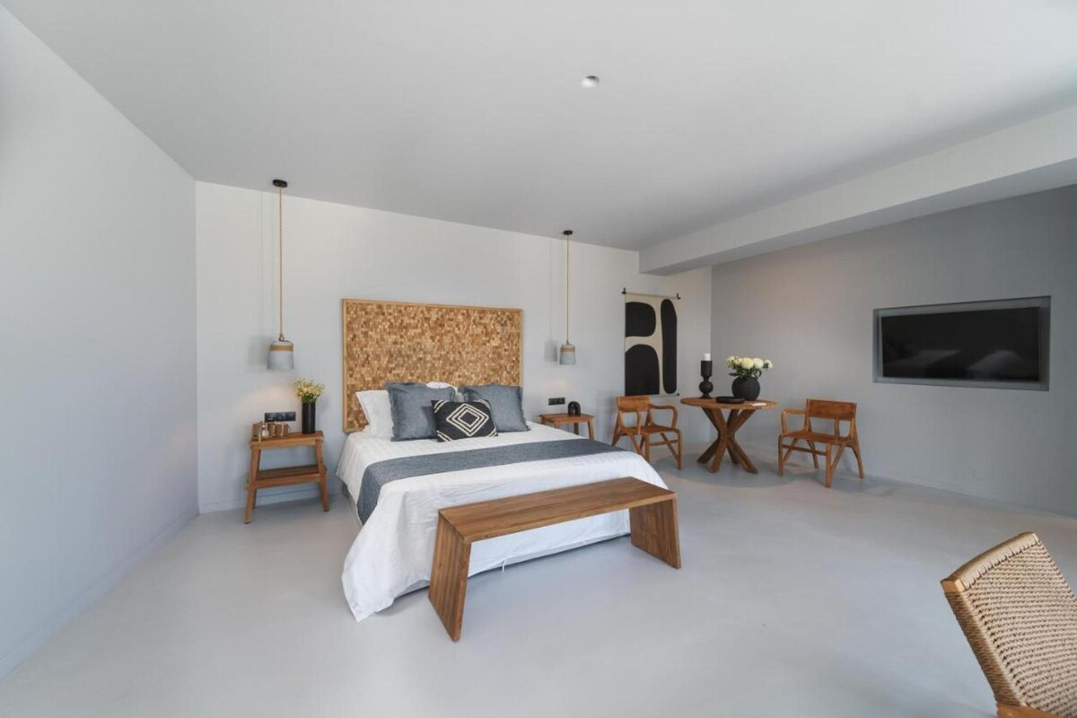 A modern bedroom with a large bed, bedside tables, a wooden bench, a small round table with chairs, and a TV mounted on the wall.