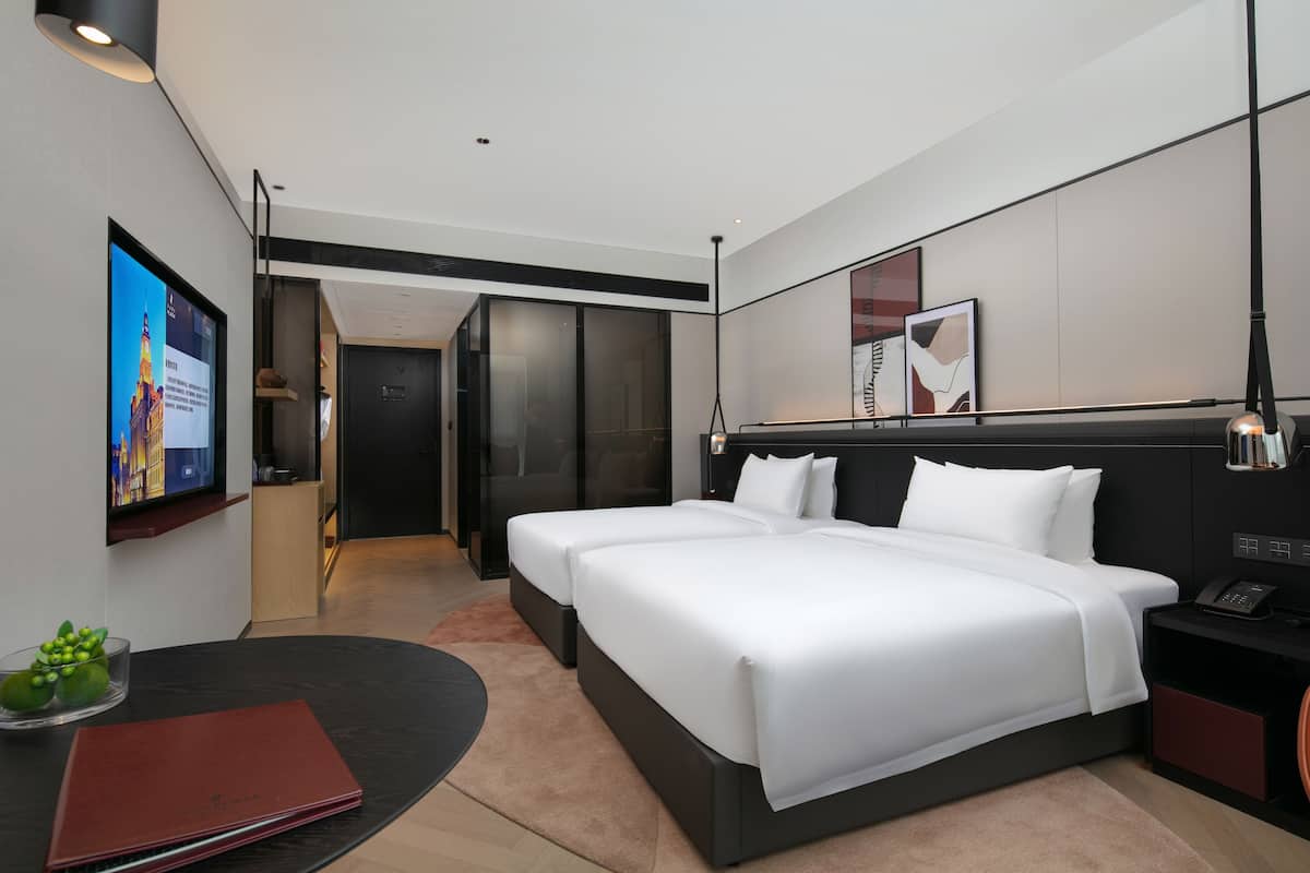Ein modernes Hotelzimmer mit zwei Betten, einem Fernseher, einem Schreibtisch und einer kleinen Pflanze.