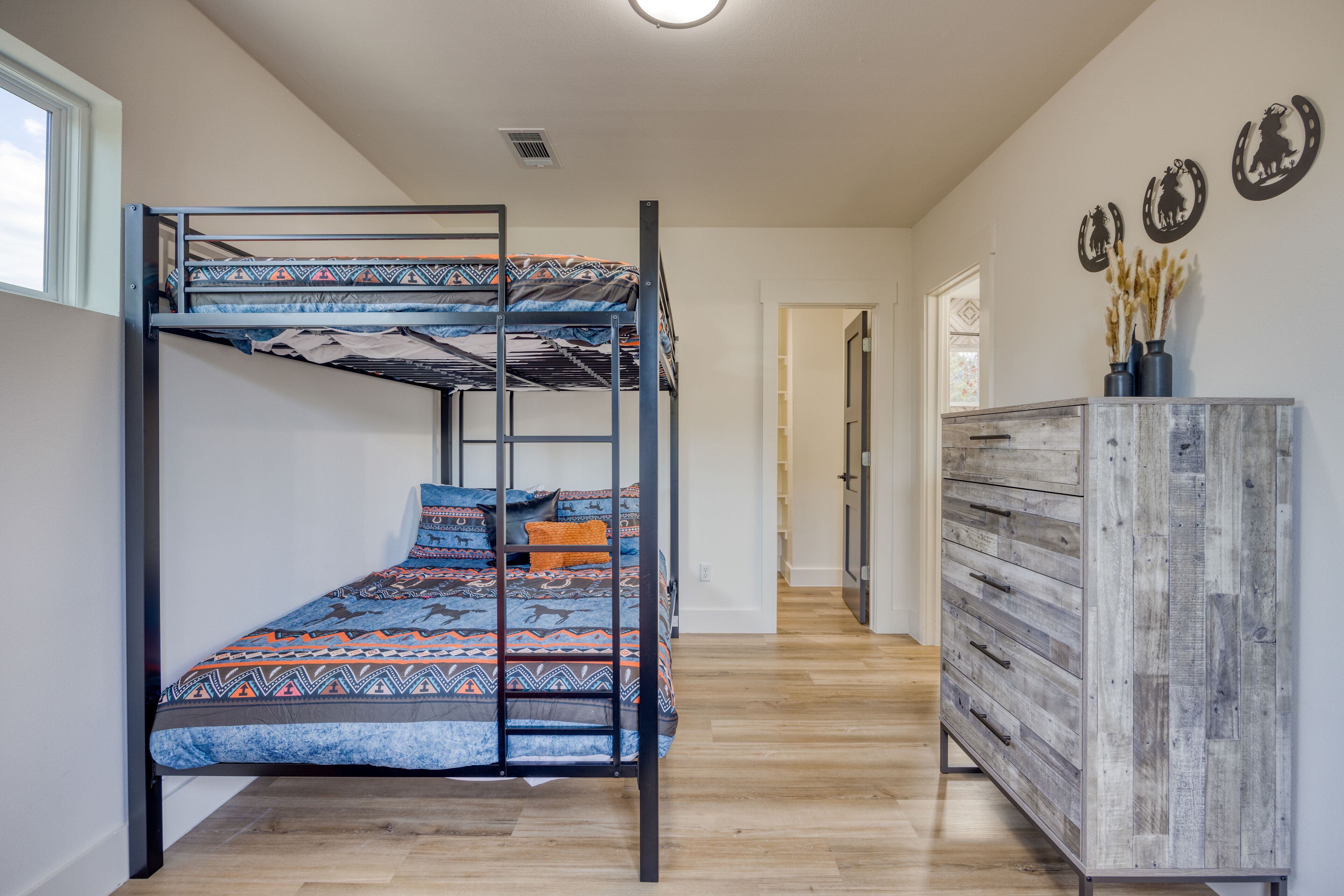 A bunk bed with patterned bedding, a wooden dresser, and decorative wall art in a room with wooden flooring and a window.
