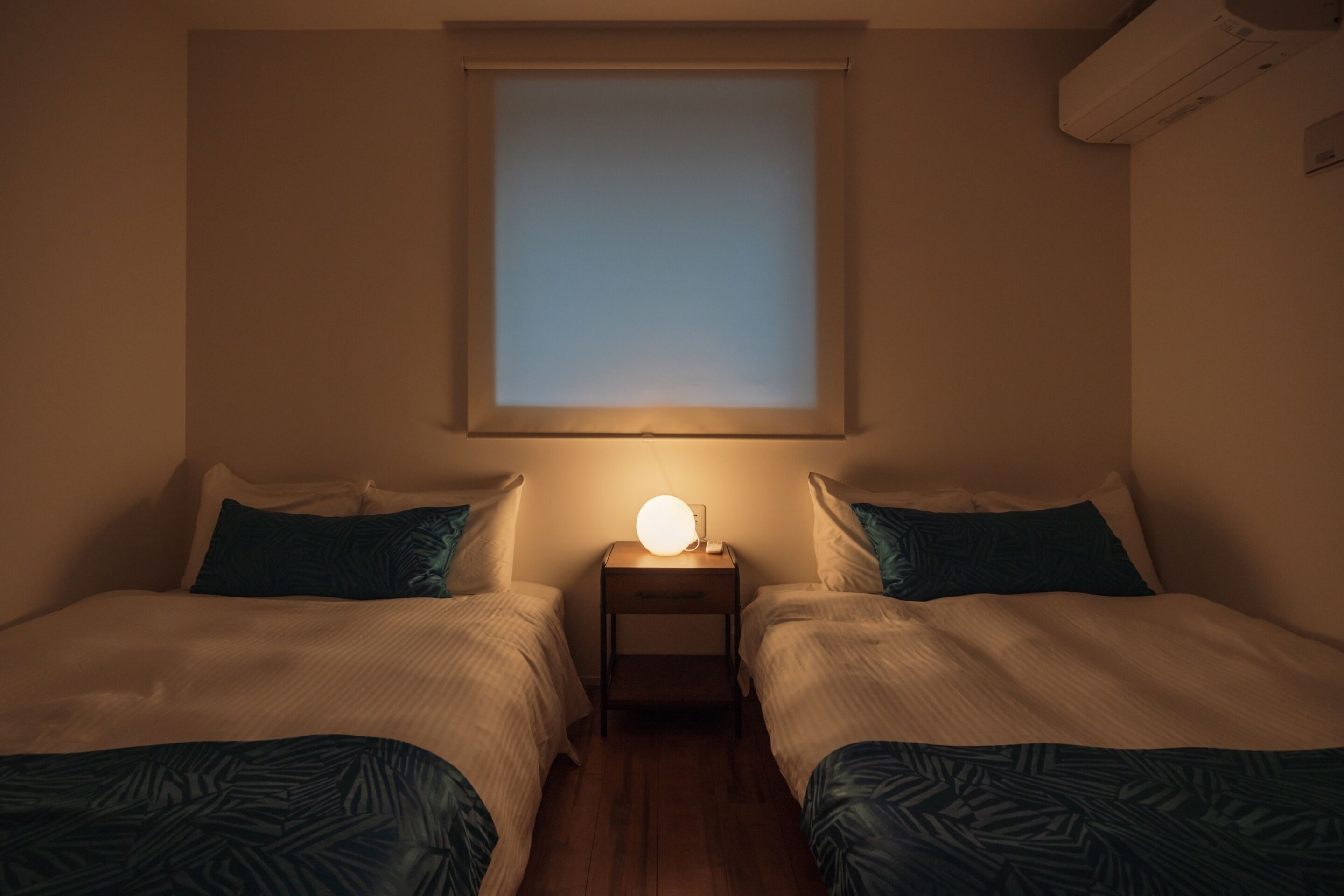 A bedroom with two beds, a bedside table with a lamp, a window, and an air conditioning unit.