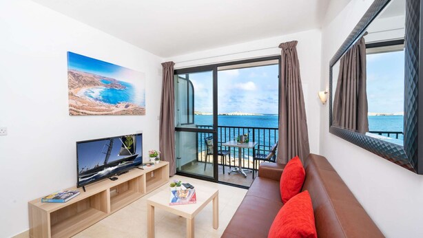 A modern living room with a large TV, a sofa, a small table, and a balcony with a view of the sea.