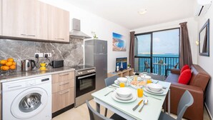 A modern kitchen with a washing machine, a dining area with a table set for breakfast, and a balcony with a view of the sea.