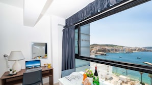 A hotel room with a desk, laptop, and a dining area with table set for two. A large window offers a view of a coastal city and the sea. - Islet Promenade Seaview Studio (Bugibba)