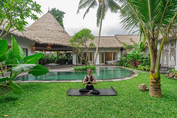 A person meditating on a mat in a lush garden with a pool and thatched-roof buildings.