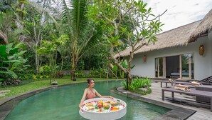 Uma área à beira da piscina com mesa de jantar, cadeiras de descanso e um edifício com telhado de palha.