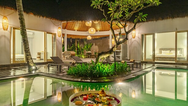 Uma área de piscina com espaço para refeições, espreguiçadeiras e uma estrutura com telhado de palha.