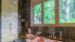Uma banheira cheia de pétalas de flores, um chuveiro e uma janela com vista para o verde.