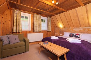 Una habitación acogedora con paredes de madera, una cama con ropa de cama morada, un sofá verde con almohadones decorativos y una mesa de madera.