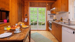 A wooden cabin interior with a dining table set for breakfast, a kitchen with modern appliances, and a large glass door leading to a green outdoor area. - Waldhaus: Sauna Jacuzzi Kamin 16 Pax (Alpirsbach)
