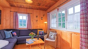 A cozy cabin interior with a wooden ceiling, a sofa, a wicker chair, and a table with a vase of flowers and a wine glass.