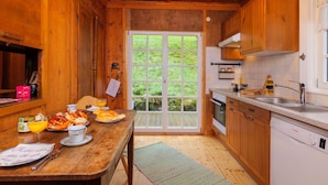 A wooden cabin interior with a dining table set for breakfast, a kitchen with modern appliances, and a large glass door leading to a green outdoor area.