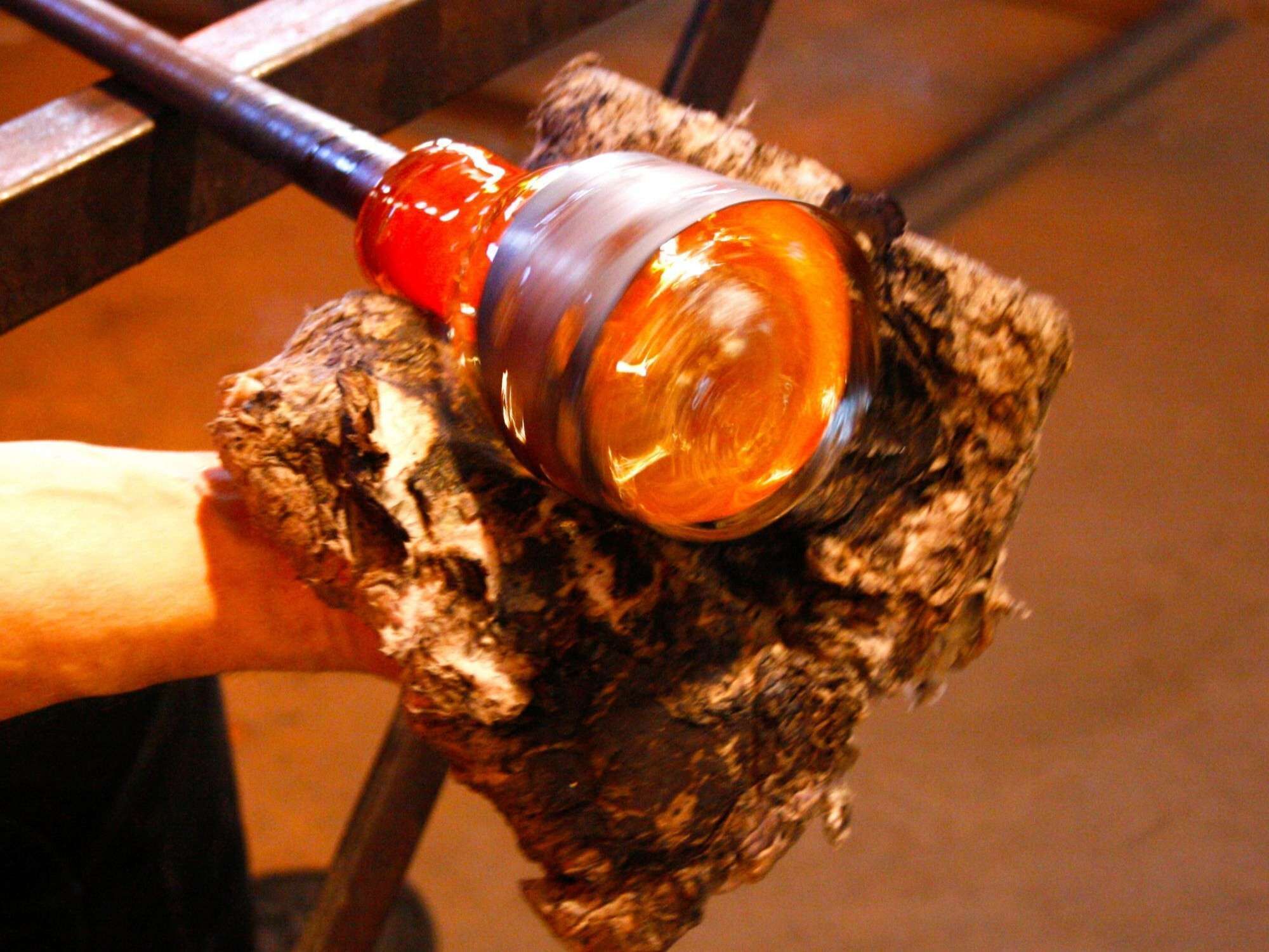 A hand holding a glass blower's tool, shaping a glass object.